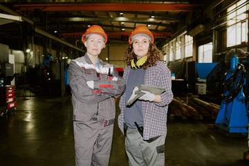 Obraz premium Two Caucasian women standing in industrial factory wearing protective helmets, one crossing arms while other holding gloves and clipboard, both looking confidently at camera