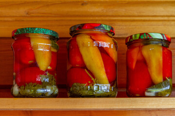 Homemade pickled peppers with tomatoes in glass jars on a wooden shelf.