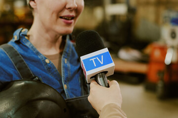 Caucasian middle aged woman being interviewed by reporter holding microphone with TV logo, standing in industrial setting, wearing work overalls, smiling and speaking