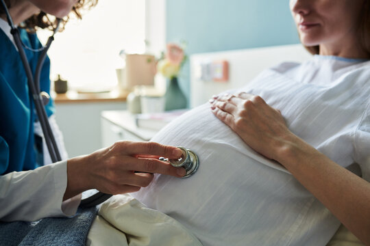 Caucasian young adult woman lying in hospital bed receiving prenatal checkup from female healthcare professional using stethoscope on pregnant belly during medical examination - Powered by Adobe