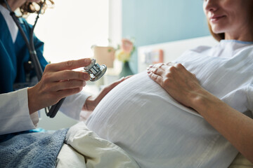 Caucasian pregnant woman sitting during medical examination with female doctor holding stethoscope near belly, focusing on prenatal healthcare and pregnancy monitoring