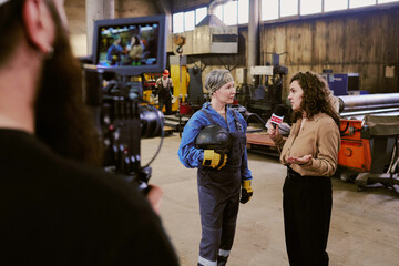 Caucasian middle aged woman in work overalls holding helmet being interviewed by young adult Caucasian woman with microphone while cameraman filming scene in industrial workshop
