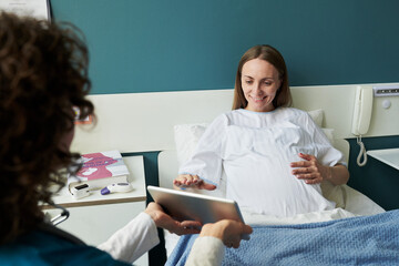Pregnant Caucasian young adult woman lying in hospital bed smiling and touching belly while female doctor showing information on digital tablet during prenatal consultation