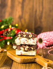 Stack of festive chocolate and white chocolate peppermint bark with hazelnuts on a wooden board, set on a rustic table with Christmas decorations and red twine in the background