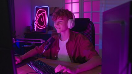 Zoom in view of excited teenage streamer speaking into professional microphone celebrating winning while recording gameplay at wooden desk in neon lit room