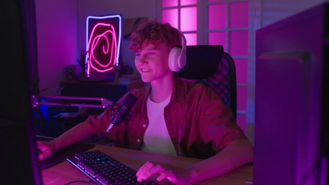 Zoom in view of excited teenage streamer speaking into professional microphone celebrating winning while recording gameplay at wooden desk in neon lit room