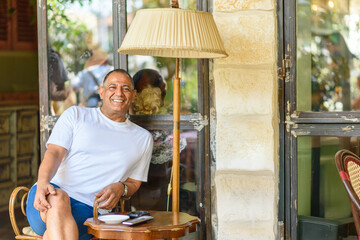 A smiling man sits casually by a lamp in a outdoor cafe, basking in the warm sunlight while enjoying a relaxing moment, surrounded by a friendly atmosphere and people chatting nearby.