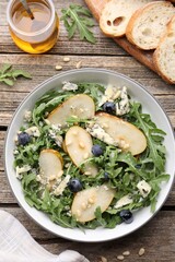 Tasty salad with arugula, pear, blue cheese, peanuts and blueberries in bowl on wooden table, flat lay