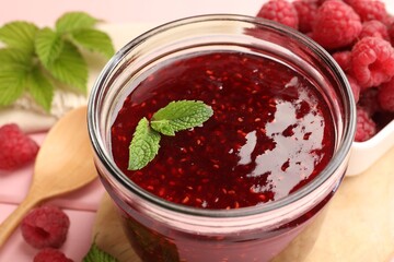 Sweet raspberry jam in glass jar, berries and mint on table, closeup