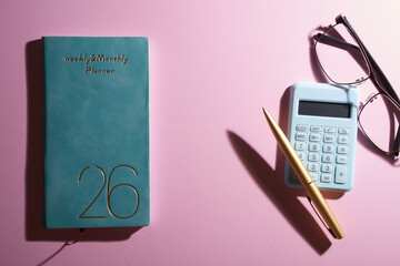 2026 small planner calculator, eyeglasses and pen against pink background.