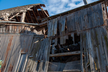 A dramatic, low-angle view of an old, deteriorating barn, showing the collapsed roof and heavily damaged wooden walls with a bright blue sky above.