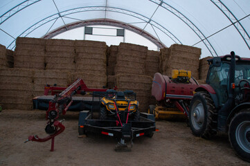 A wide view of various pieces of farming machinery, including a tractor, ATV, and baler, stored next to stacked hay bales inside a hoop barn.