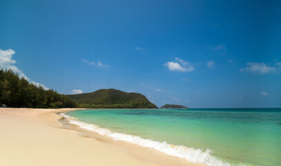 Peaceful tranquil tropical sea beach with crystal clear turquoise water gently washing onto soft sandy shore, framed by lush green hills and bright blue sky dotted with fluffy white cloud for travel