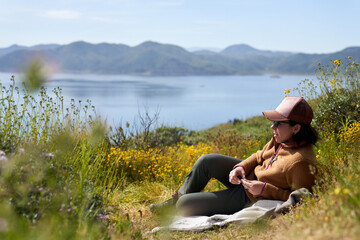 A girl in a brown jacket and hat rests in a field overlooking water