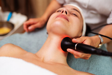 Beautician performing radiofrequency lifting procedure on female patient's neck in beauty salon,...