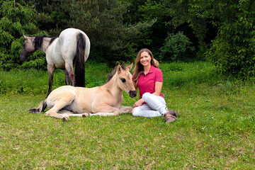Young woman sitting on ground next to dunn color small horse foal, another animal near in background © Lubo Ivanko