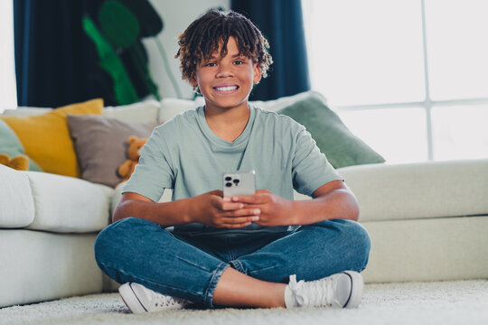 Charming teenage boy smiling at home while using a smartphone on a cozy living room floor during leisure time