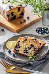 Pieces of homemade blueberry lemon cake on a plate on a light background with fresh berries, herbs and morning light. Summer healthy dessert concept. Top view.