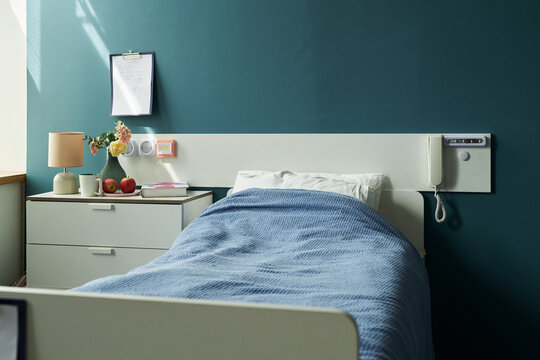 Empty hospital bed with blue blanket standing in clean patient room, bedside table holding lamp, flowers, fruit and clipboard hanging on wall, medical phone attached to headboard