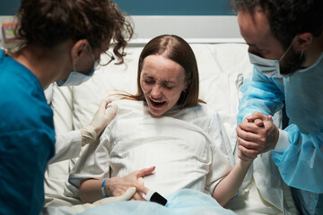 Caucasian young adult woman experiencing labor pain in hospital bed, supported by medical professional and Caucasian young adult man holding her hand during childbirth process