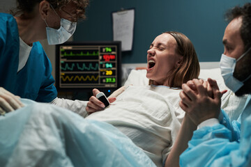 Caucasian young adult woman experiencing labor pain in hospital bed surrounded by medical staff and partner, gripping hand tightly while undergoing childbirth process