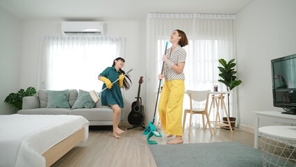 Fototapeta premium Caucasian mom and asian child doing housework together and dancing to music. Attractive mother and daughter cleaning modern bedroom while sing along and jump to relaxing music. Lifestyle. Pedagogy.