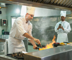 professional chef in white uniform action skillfully cooking in industrial kitchen on stovetop with visible flames, while another chef captures moment on smartphone in modern, well-equipped kitchen.