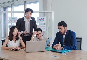 Group business professionals team having meeting in modern office setting discussing work around table with laptop notebook and smartphone. Charts and graphs can be seen on glass wall behind them.