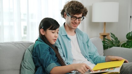 Happy daughter lean on father shoulder while listening story telling. Young asian schoolgirl looking ar dad while reading story book together and sitting at sofa. Family recreation concept. Pedagogy.