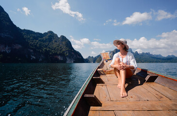 A woman sitting on a traditional wooden boat, enjoying the serene lake scenery surrounded by lush green mountains under a sunny sky, offering a moment of tranquility and connection with nature.