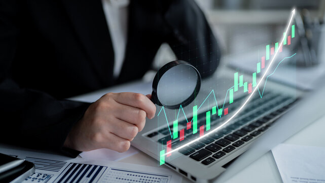 A business professional uses a magnifying glass to analyze stock market trends displayed on a laptop, symbolizing financial growth and market insight. Impute - Powered by Adobe