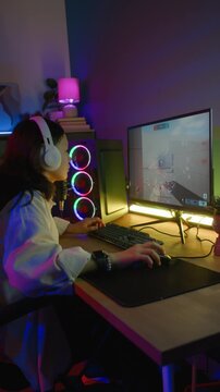 Vertical shot of female teenage gamer with headphones playing third-person shooter on computer in cozy room with neon lighting