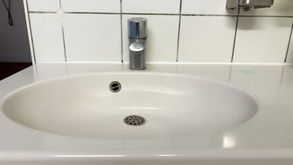 Clean white oval bathroom sink with chrome faucet, close-up view of ceramic basin and metal drain on tiled wall background, minimalist washroom interior design and hygiene concept