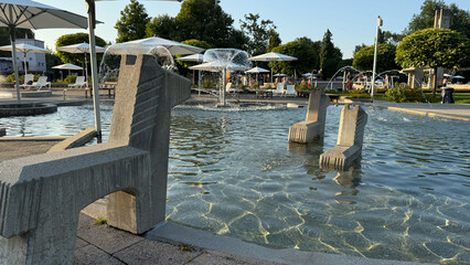 Swimming pool with decorative fountains at sunset, outdoor public recreation area with parasols and water features, summer leisure scene, tranquil atmosphere, vacation relaxation concept