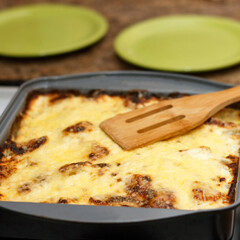 Baked lasagna in a black tray on a stove. A wooden spatula lies on top