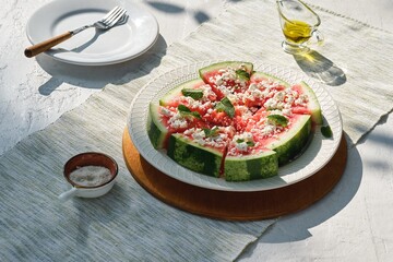 Appetizer, watermelon chunks with rind, sprinkled with crumbled feta cheese, drizzled with olive oil and garnished with mint leaves on a ceramic plate.