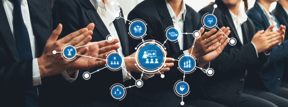 A group of business professionals in suits applaud while a digital overlay shows various icons representing collaboration, success, and innovation in a corporate seating arrangement. Amity - Powered by Adobe