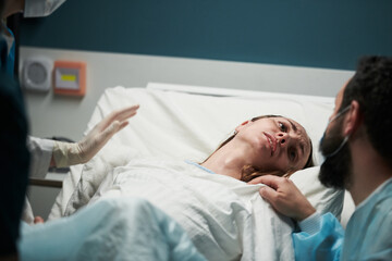 Caucasian young adult woman lying in hospital bed experiencing labor pain, looking distressed while...
