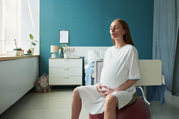Caucasian pregnant young adult woman sitting on exercise ball practicing breathing techniques in hospital room hands resting on belly preparing for childbirth