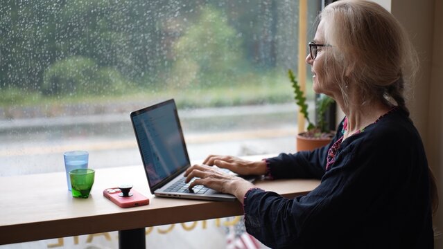 A smiling mature woman sits at a cafe window, sipping coffee and multitasking on her laptop and phone while rain falls outside. She appears happy and content.
