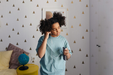 Black girl listening to music on smartphone with earphones in bedroom