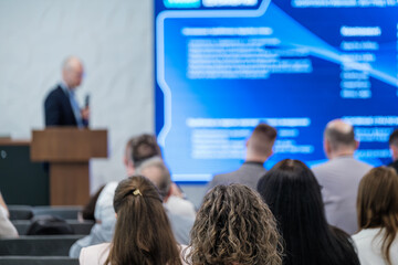 Conference speaker presenting to attentive audience with focus on speaker presentation and screen.