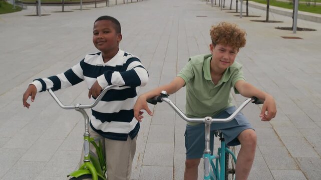 Tilt up of two cute smiling boys in casual attire sitting on their bicycles and looking at camera while standing on wide sidewalk with rows of trees growing in park