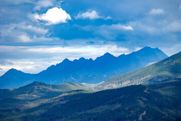 Dramatic mountain silhouette under cloudy blue sky with sunlit forested hills