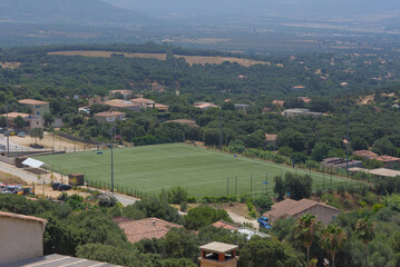 An aerial view shows a green soccer field surrounded by a residential area with houses and trees, with a vast, forested landscape in the background. © Marc