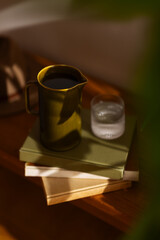 Close-up of a ceramic jug filled with water and a glass of water on stacked books in warm natural light. Cozy and minimal home setting with soft shadows.