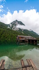 Naklejka premium A wooden boathouse sits on a clear, green lake, with a mountain shrouded in clouds in the background.