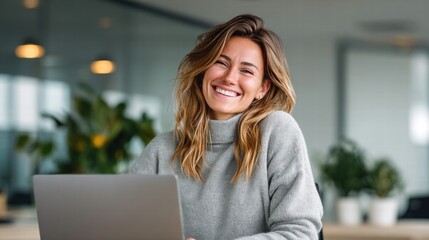 Happy woman smiling brightly while using a laptop at her workplace, looking at the camera.