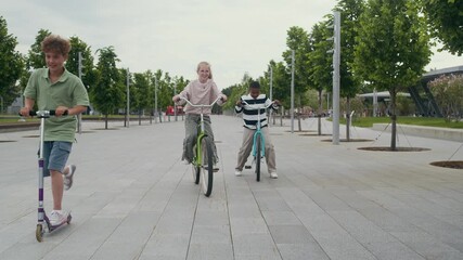 Group of four friendly diverse children wearing casual attire riding bicycles and scooters along sidewalk between two rows of green trees growing in park