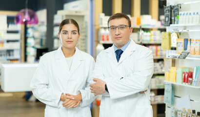 Obraz premium Two smiling female and male pharmacist workers stand shoulder to shoulder and affably welcoming visitors in pharmacy s sales area